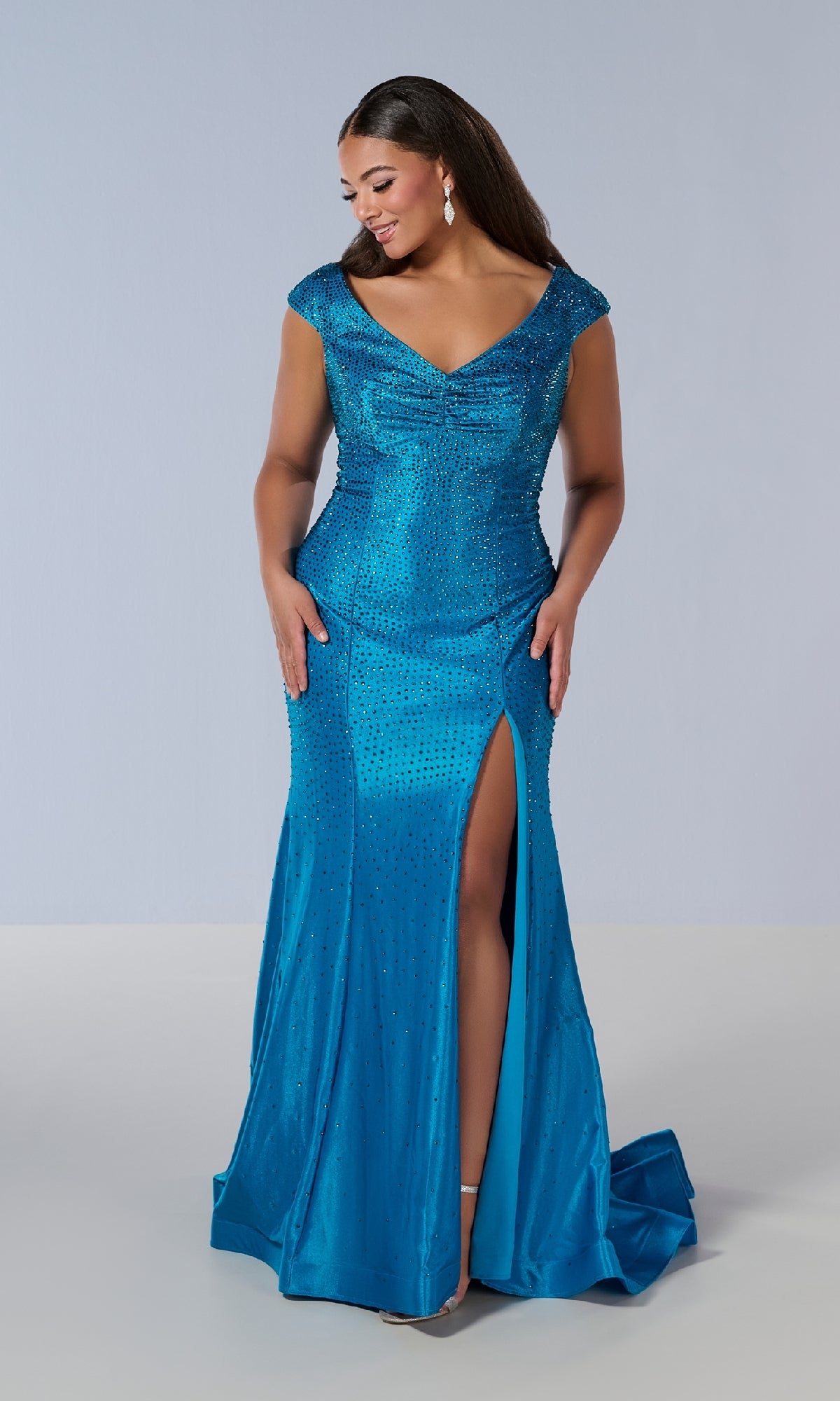 A woman in the Tiffany 16198 long plus-size prom dress—fitted, floor-length, blue, with cap sleeves, a V-neckline, and a thigh-high slit—stands against a plain background, looking down and smiling.
